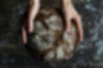 Two hands holding a round, crusty loaf of artisan bread against a dark background.
