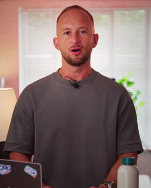 Person smiling, wearing a gray shirt, with a mic headset. Background has window blinds and a plant. Laptop with stickers and a bottle visible.