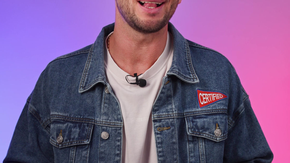Smiling man in denim jacket with "CERTIFIED" patch, against a pink-to-blue gradient background. He holds hands in mid-action, with a microphone clipped on.