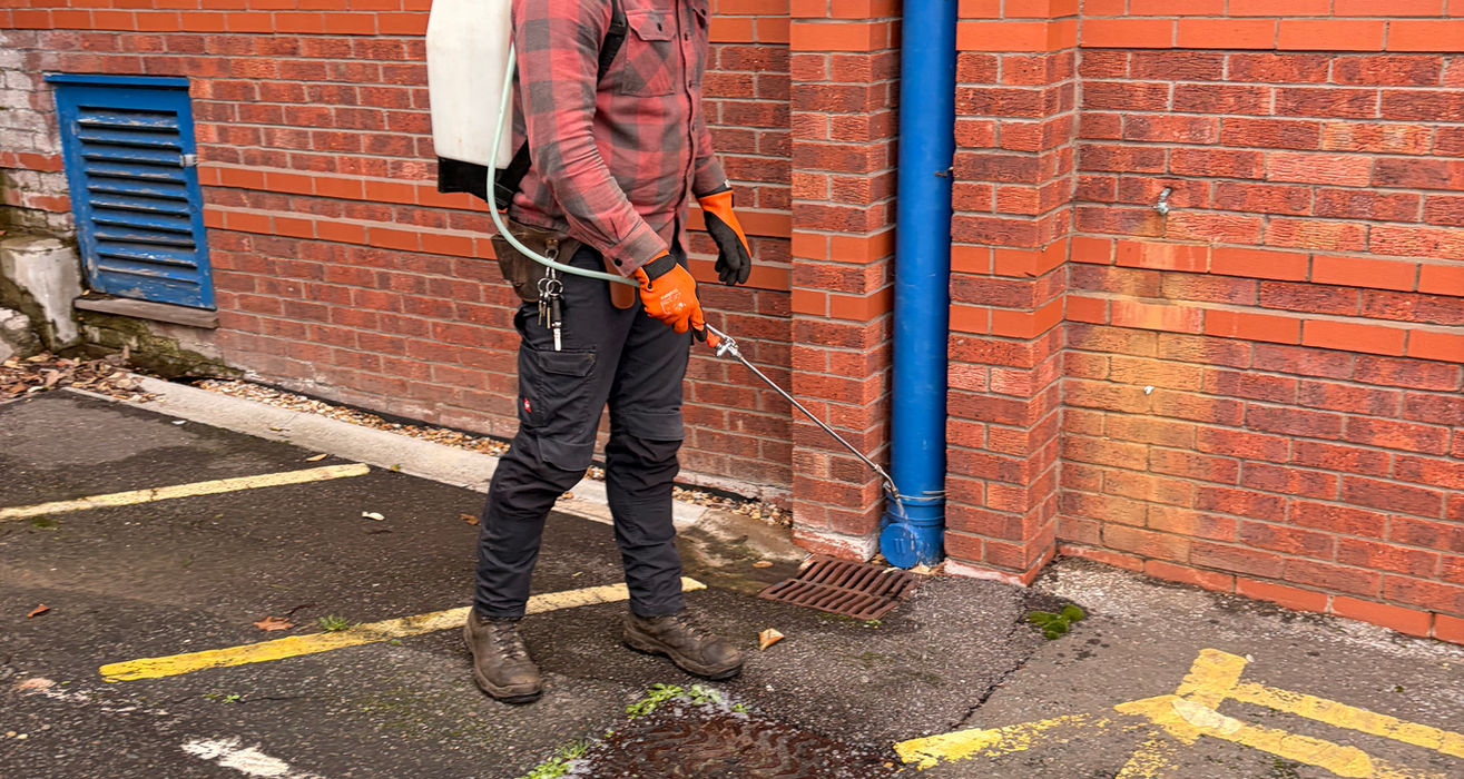 Man with backpack sprayer treats weeds in industrial parking lot.