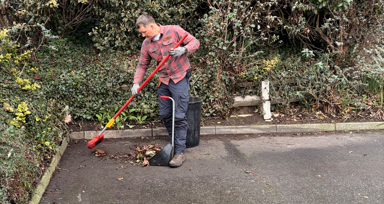Man raking leaves with a red rake on a paved area.
