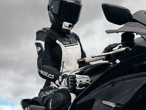 Motorcyclist in black and white gear, seated on a black bike. Overcast sky. Visible text: "Held." Calm, focused mood.
