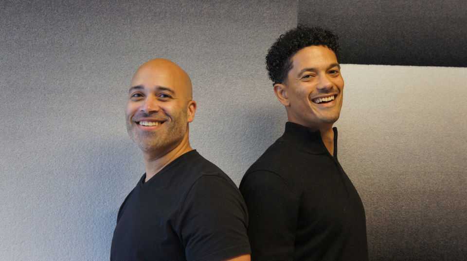 Two men smiling at the camera, standing back-to-back, wearing black shirts