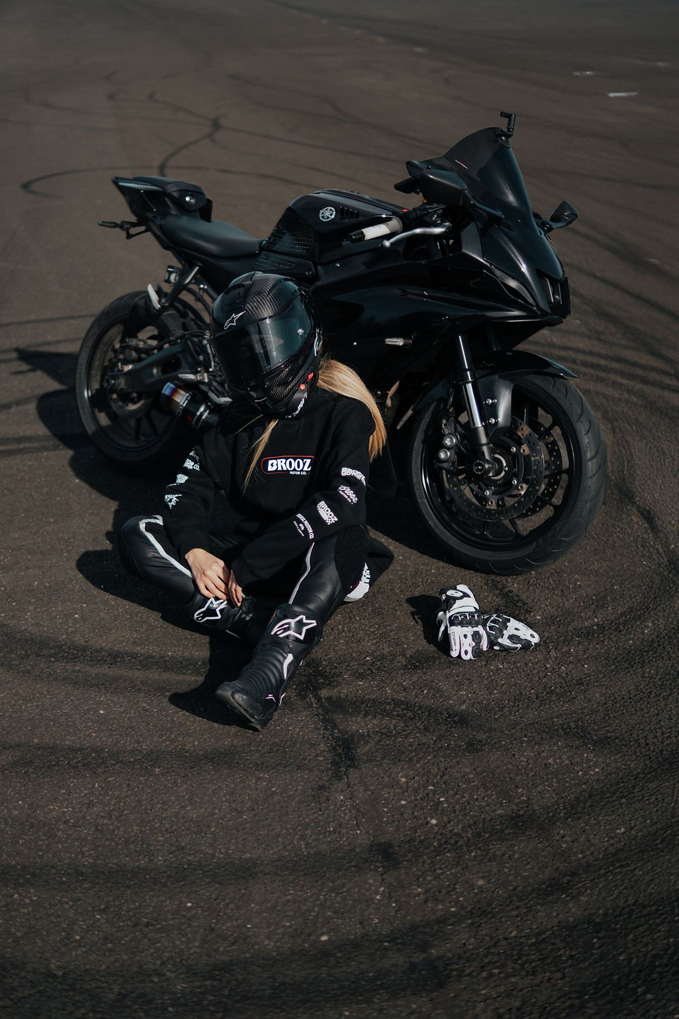 A person in motorcycle gear sits on asphalt beside a black bike. Black and white gloves lay nearby. The scene is calm, with visible tire marks.