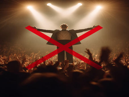 A stage hypnotist, wear a tuxedo, on stage with his arms spread out to a large crowd with a large red X over him