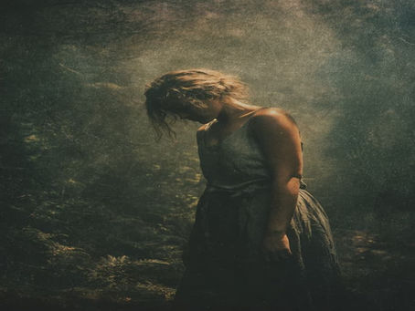 A sad woman with standing with her head down surrounded by dark light with dim light shining on her, symbolizing feeling lost after a breakup