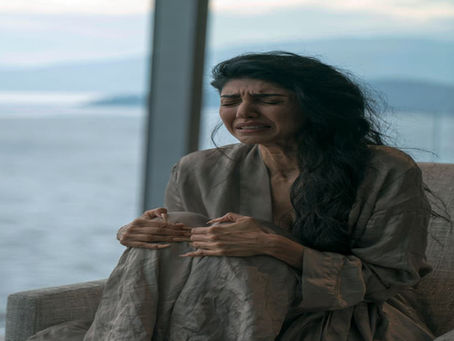 Persian woman in a silk robe grips her knee in pain, face contorted, seated by a window with a water and mountain view in her West Vancouver home