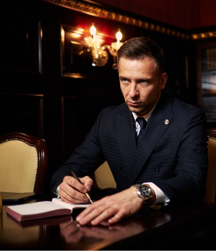 Erkek, şık takım elbise içinde, karanlık ahşap bir ofiste masa başında ciddi bir ifadeyle deftere yazıyor. Arka planda avize ışıkları.