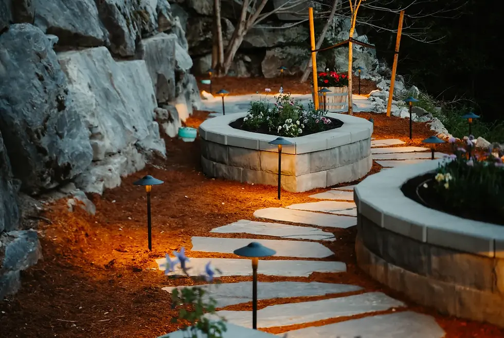 Lit garden path with curved stone planters, colorful flowers, and rock wall backdrop. Warm lights create a calm, inviting atmosphere.
