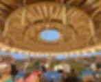Crowd seated inside a large circular wooden pavilion at dusk, watching a powwow with flags and chairs on grassy ground.
