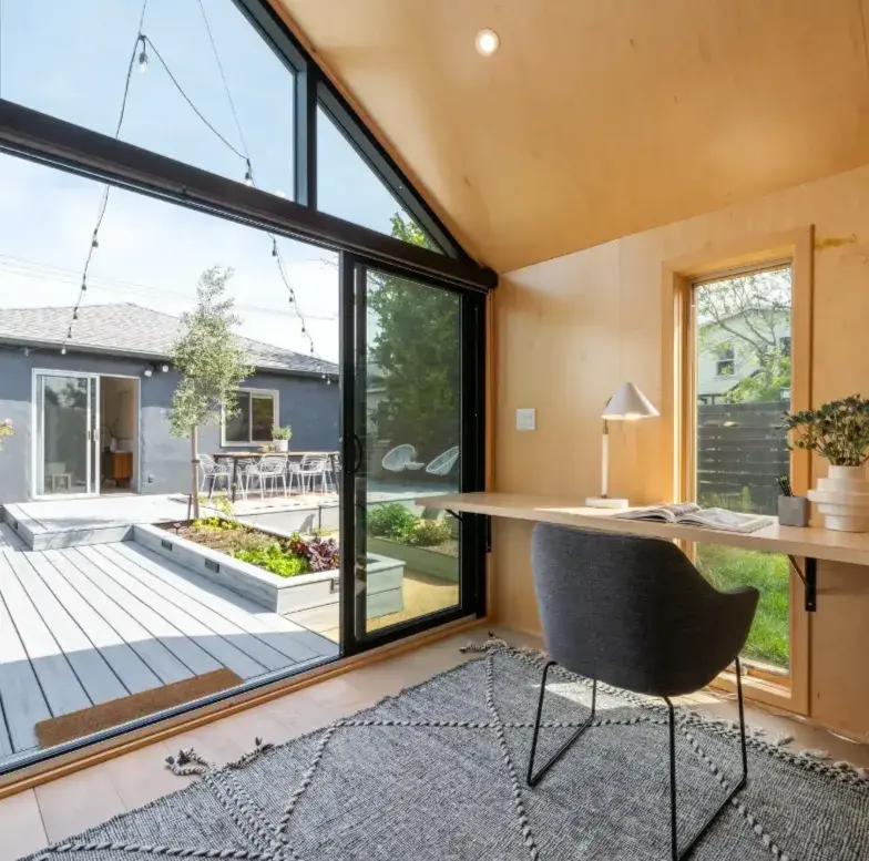 Modern home office with a large window showing a patio garden and gray house. Inside, a desk with lamp, chair, and open book on a gray rug.