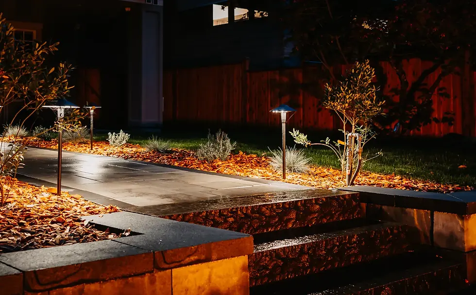 A dimly lit stone pathway at night with steps, bordered by mulch and small plants. Path lights cast a warm glow. Wooden fence in background.