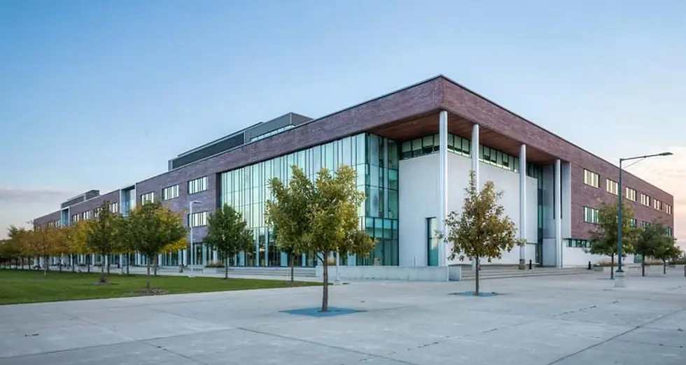 Modern building with glass facade and brick exterior. Trees line the front on a wide concrete path. Clear sky creates a calm mood.