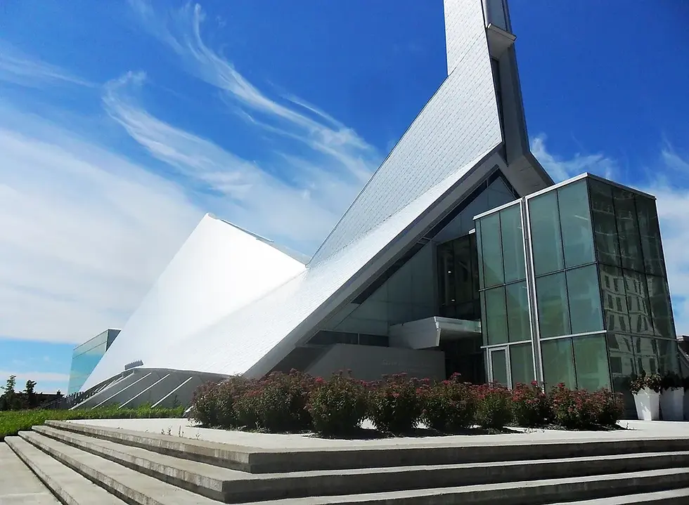 Modern building with sharp angles and glass facade under a bright blue sky. Steps lead up to the entrance, with shrubs in front.