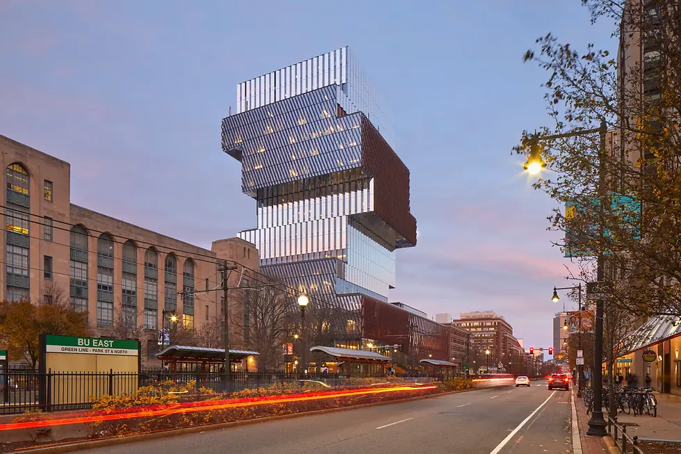 Modern stacked building with glass facade at dusk, streetlights on. BU East sign visible, trees line the street, light trails from cars.