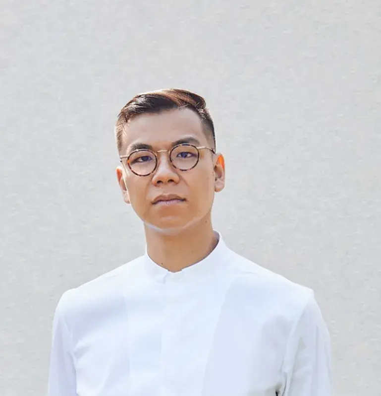 Man in glasses and white shirt stands against a plain background, looking confidently at the camera. The mood is calm and composed.