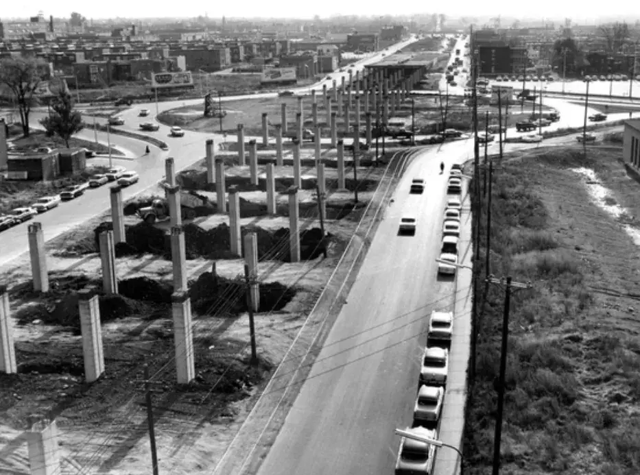 Construction site with rows of concrete pillars next to a road lined with cars. Urban landscape in the background. Black and white photo.