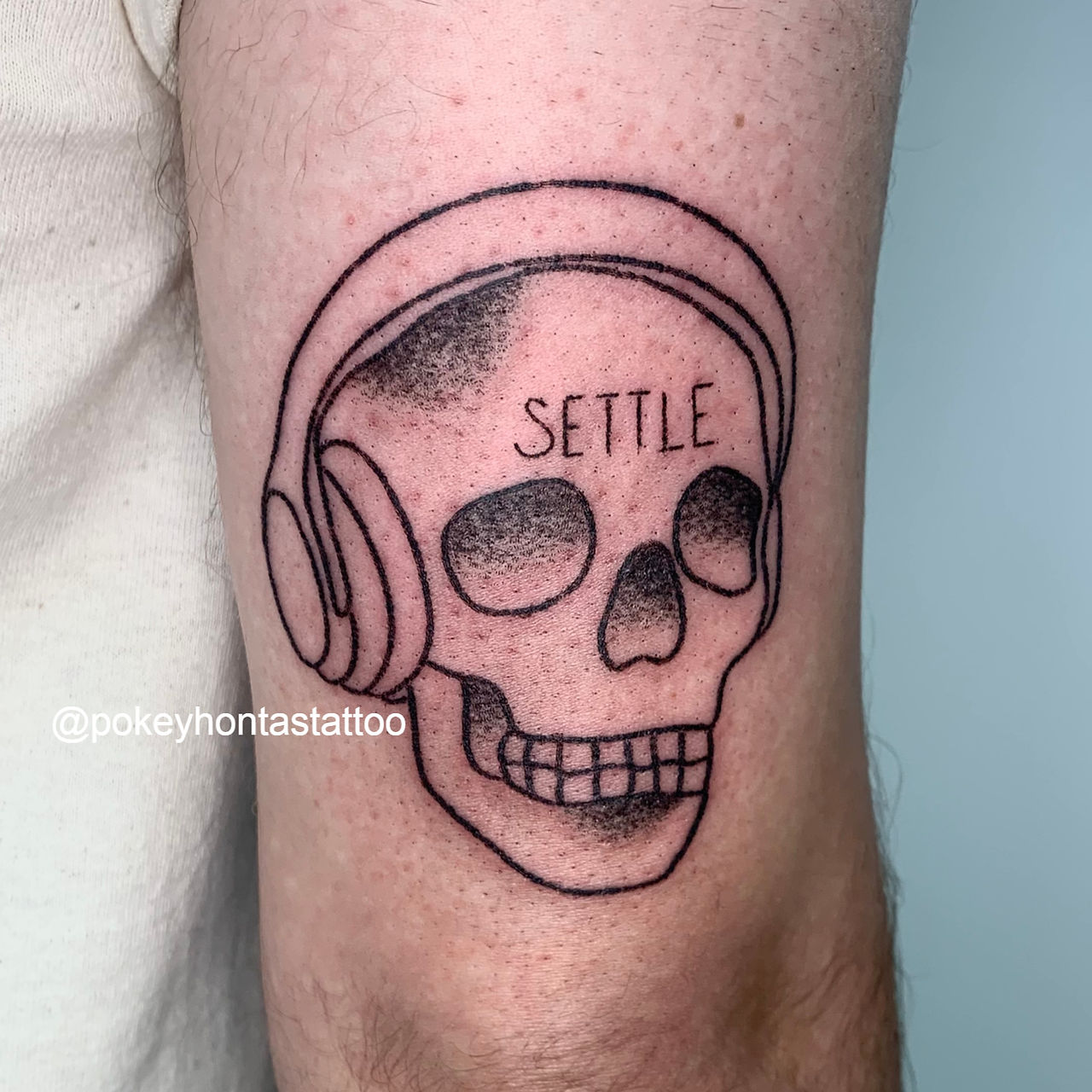 A stick and poke tattoo of a skull wearing a pair of headphones on the back of the right upper arm