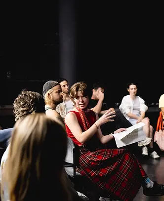 Izzy sitting in a group of people, dressed in red, explaining something | Izzy Collie-Cousins Theatre Director