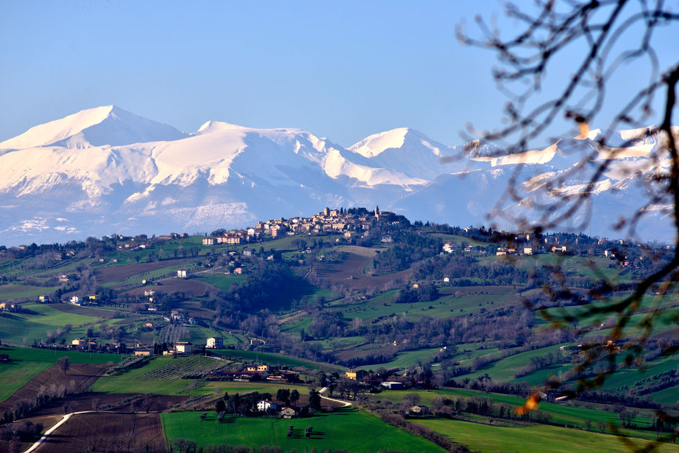 Paesaggio collinare con un borgo in cima a una collina e, sullo sfondo, alte montagne innevate illuminate dalla luce limpida di marzo.