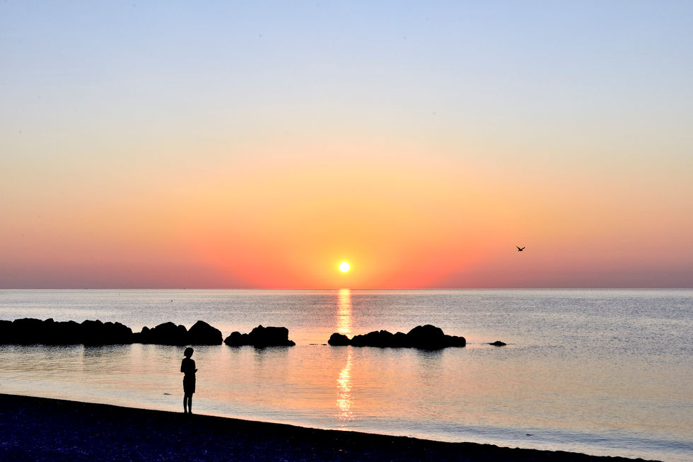 Una figura femminile minuta, immobile in riva al mare, guarda l’orizzonte all’alba. Il mare è calmo come una tavola. In alto a destra, un gabbiano in volo attraversa il cielo. La scena è bilanciata, silenziosa, sospesa. È un momento di ascolto e presenza.