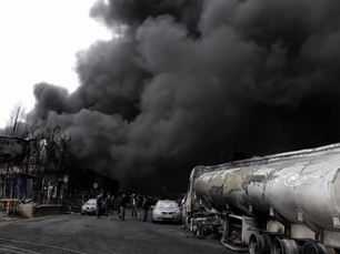 Thick black smoke engulfs tanker trucks and people on a road in Iran. A burnt sign is visible. The scene is tense and gloomy.