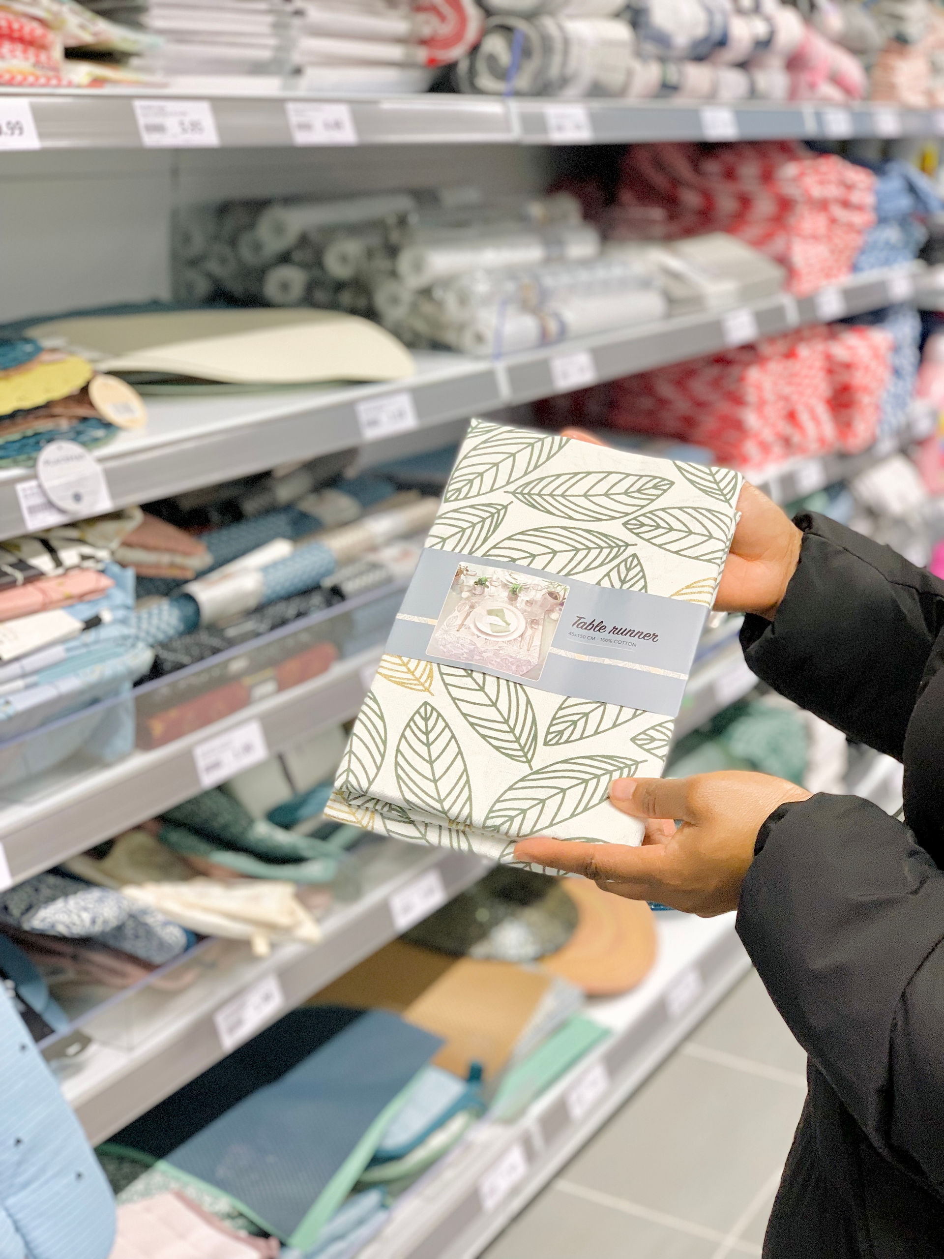 Mains d'une femme, décoratrice d'intérieure, dans une allée d'un magasin, tenant le paquet d'une nappe de table à motifs dans les tons bleu et vert.