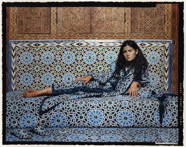 Person reclining on a patterned bench in an ornate, mosaic-tiled room, their robe and skin intricately blending with the blue, white, and green geometric Islamic-style motifs, creating a camouflaged visual effect within the richly detailed interior décor. Photograph by Lalla Essaydi