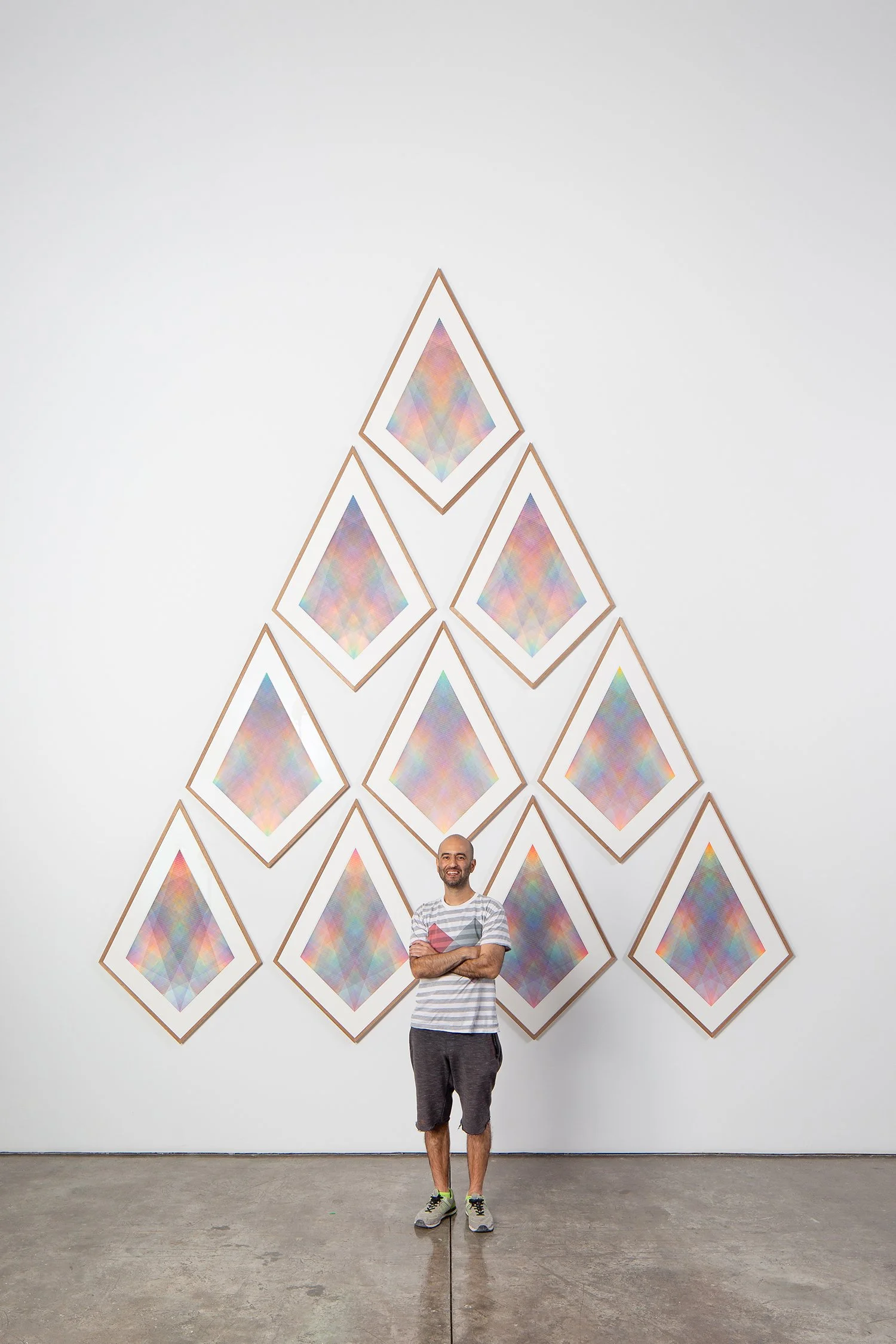 Nima Nabavi standing with arms crossed in front of a large geometric art installation composed of diamond-shaped frames arranged in a triangular pattern on a white gallery wall, each frame featuring pastel multicolor abstract patterns, modern art exhibit, contemporary geometric artwork display.