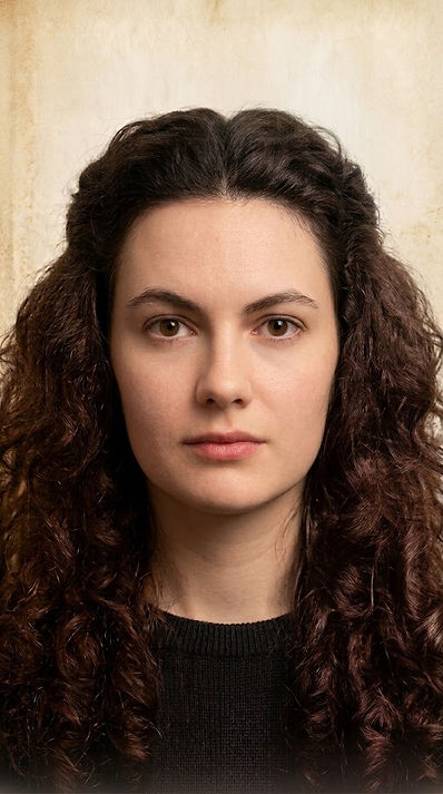 Portrait of a woman with dark curly hair and a neutral expression, wearing a black sweater against a beige background.