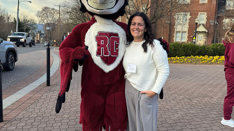 A person poses with a maroon mascot with "RC" lettering on its chest. They are outdoors on a paved path with a brick building in the background.