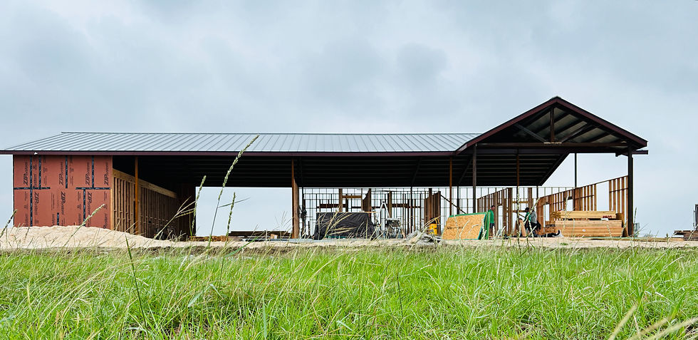 A half-built barn with a metal roof stands in a grassy field. Wooden walls and construction equipment are visible under a cloudy sky.