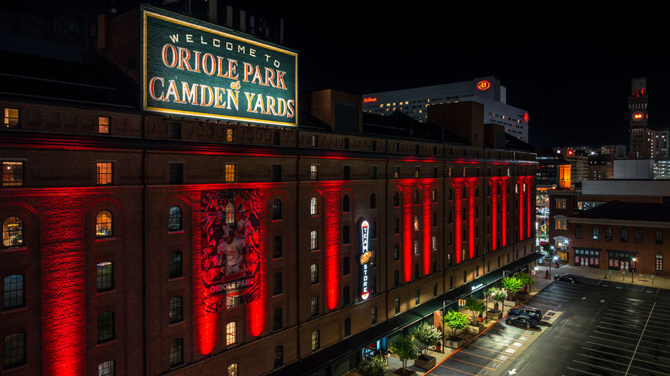 Drone photo showing Camden Yards illuminated at night with vibrant lighting in red, green, and yellow colors. The stadium lights highlight the celebration of Sickle Cell Awareness and Juneteenth, symbolizing support and recognition of both causes. The aerial view captures the iconic ballpark warehouse fully lit against the dark sky, emphasizing community unity and cultural significance.