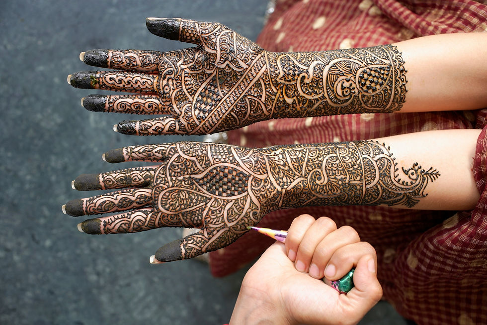 Hands adorned with intricate henna designs; an artist applies details. Background has a maroon patterned fabric, creating a traditional feel.