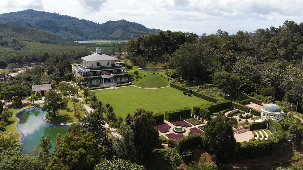Aerial view of a grand estate with a central white villa, lush gardens, a pond, and surrounding trees. Green hills and blue sky in the background; The Vanderbilt Estate