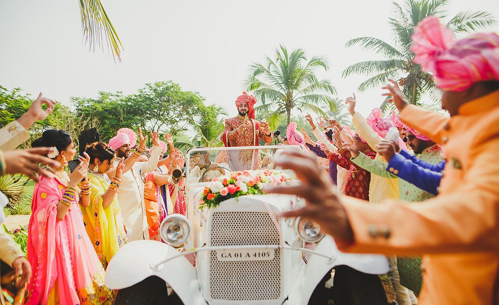 Groom in vintage car, surrounded by lively guests in colorful attire. Palm trees and bright sky in background.