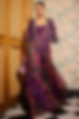 Woman in a vibrant purple floral dress and matching jacket, holding a beaded purse. Floral wallpaper and wooden door in the background.