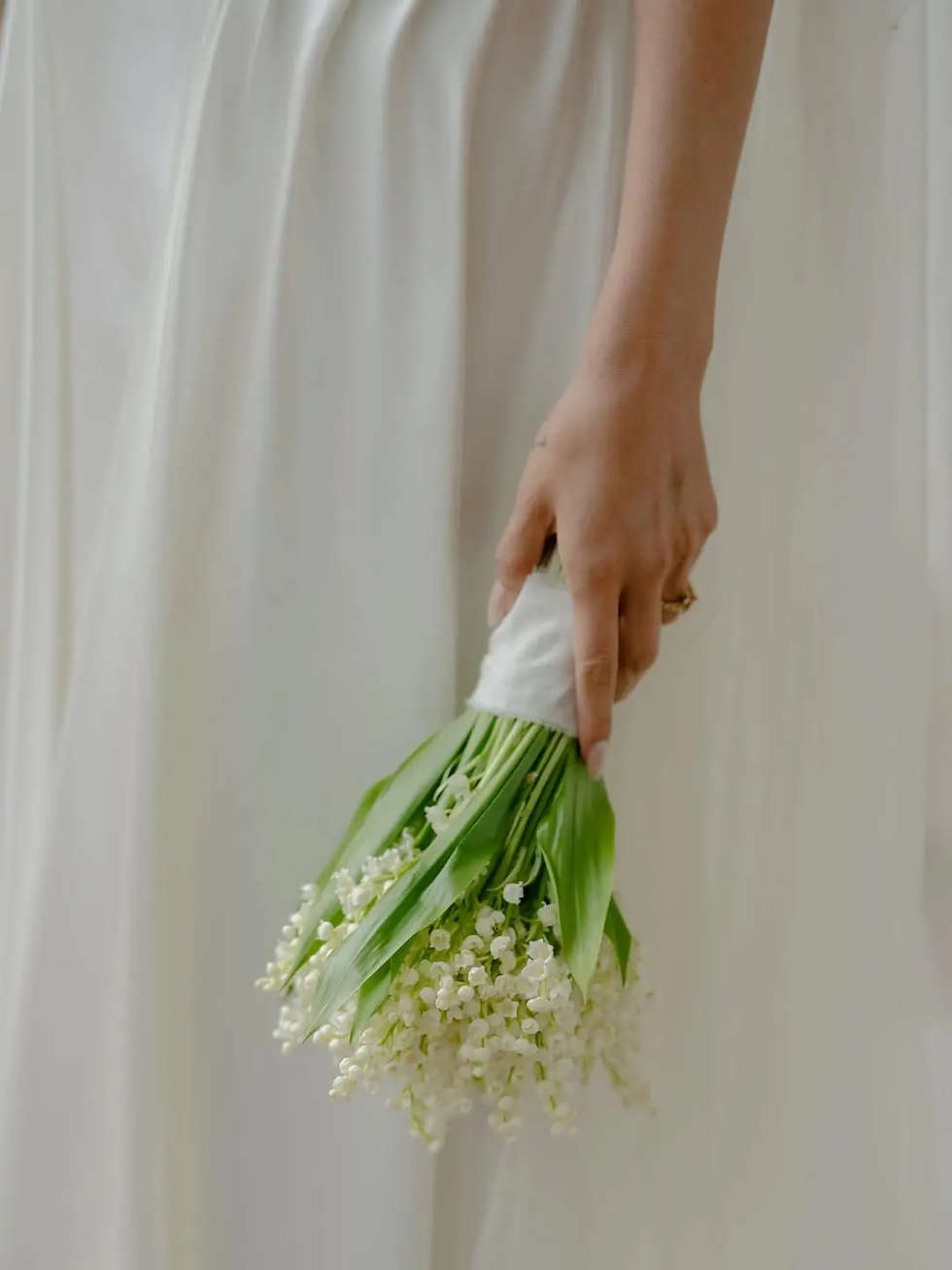 Hand holding a bunch of lily of the valley flowers against a flowing white fabric background. The scene is calm and elegant. Selena Gomez; Benny Blanco