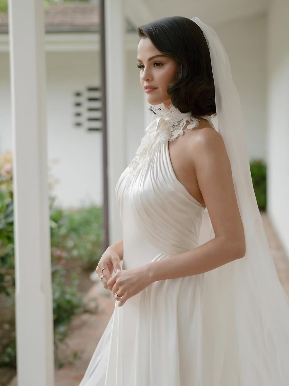 Woman in a white wedding dress with lace detail and veil stands outside, looking serene. Background features green plants and a white wall. Selena Gomez; Benny Blanco; Ralph Lauren