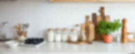 Kitchen counter display of jars with wooden lids filled with rice, flour, and sugar. Several cutting boards of various sizes are propped up and arranged against the white tile backsplash. A parsley plant is in an terracotta pot next to a wooden pepper mill grinder.