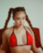 Image of Vashti Estrada sitting on a red chair in a red bikini and gold cross necklace with a jade stone.