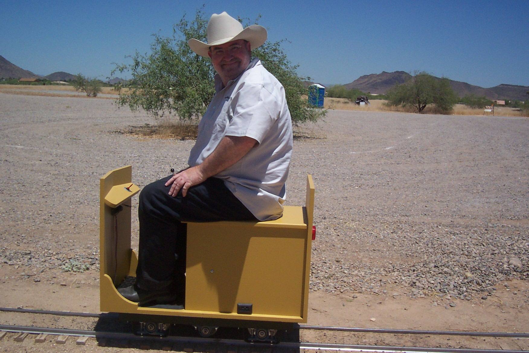Side view of a person in a white ten-gallon hat riding a yellow industrial miniature locomotive in an arid landscape
