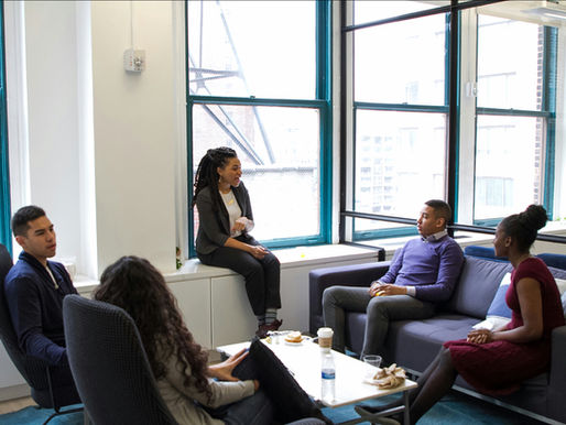 A diverse group of colleagues casually meeting and smiling in a modern office lounge with large windows. The relaxed setting emphasizes openness, inclusivity, and a collaborative workplace culture.