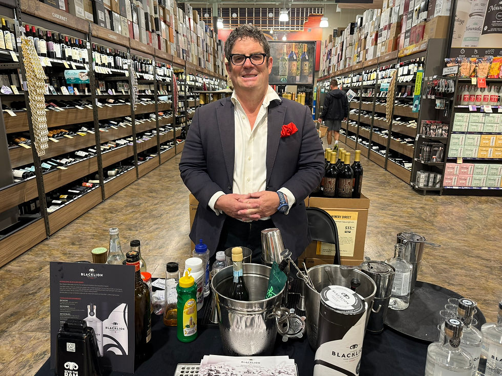Smiling man in a suit stands at a vodka tasting table in a liquor store, with rows of bottles and Blacklion promo items around him.