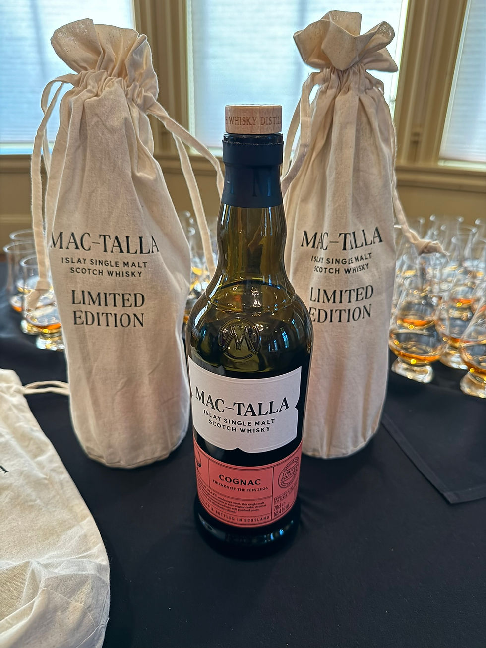 Mac-Talla limited edition Scotch whisky bottle flanked by two cloth gift bags on a table, with glassware in the background.