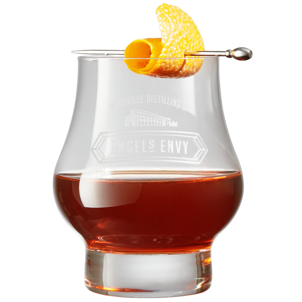 Amber whiskey in a branded Angels Envy glass, garnished with an orange peel and cocktail pick on a black background.