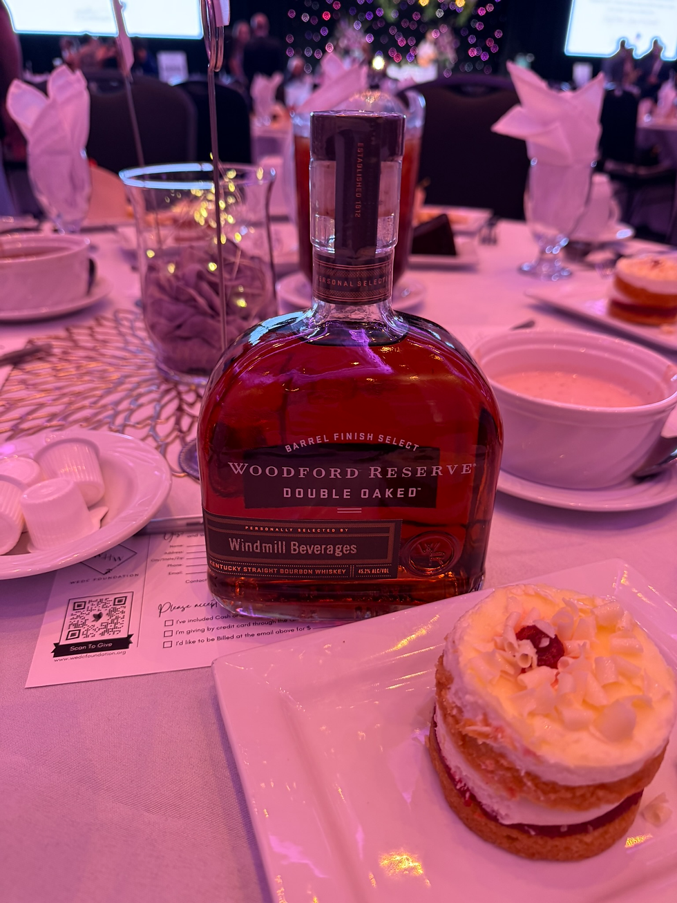 Woodford Reserve Double Oaked bourbon bottle on a banquet table beside dessert cake, under pink lighting, with place-setting cards.