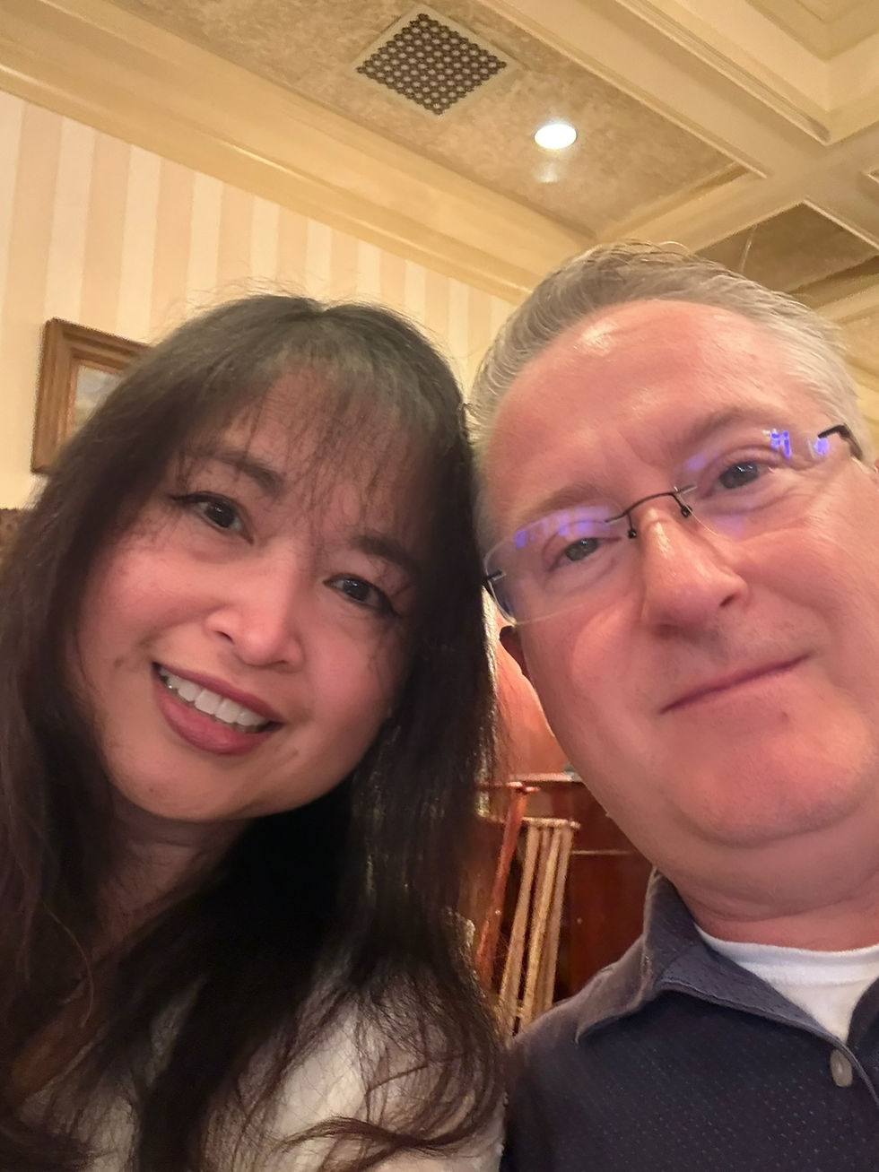 Smiling woman and man take a close selfie in a warm-lit restaurant, with striped wallpaper and ceiling lights behind them.