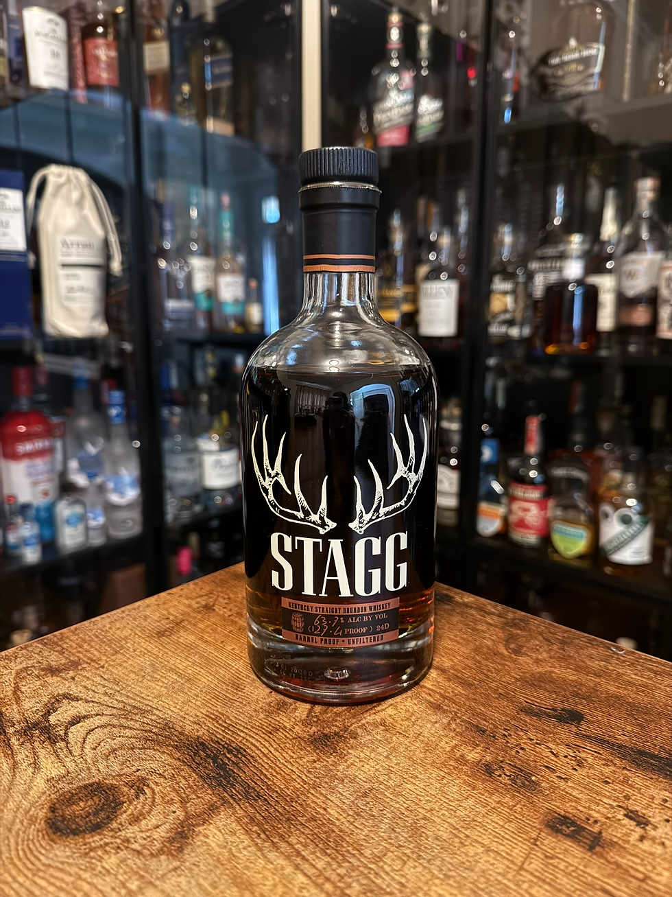 STAGG bourbon bottle with antler logo on a wooden counter, liquor shelves blurred behind, label showing Kentucky straight bourbon whiskey.