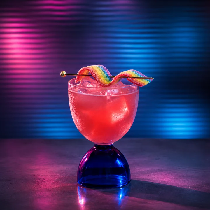 Pink cocktail with ice in a blue stemmed glass, topped with rainbow candy, against a neon pink and blue background.